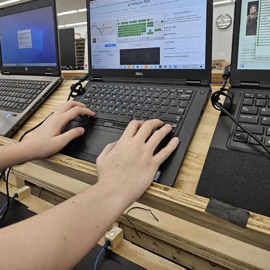 A technician’s hands on the keyboard of a refurbished laptop during testing, with more laptops cabled up along the bench.