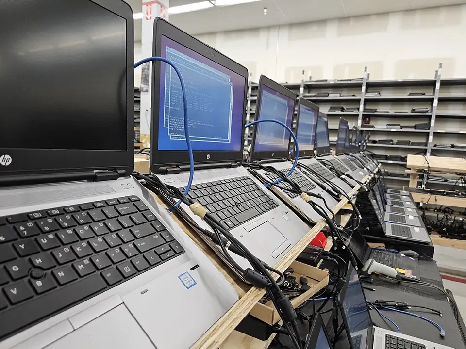A row of HP laptops open on a wooden test bench, each cabled in and running a diagnostic, with shelves of laptops behind.